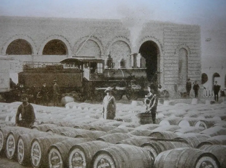 Toneles en fila en la playa de cargas de la bodega Tomba, con una locomotora detrás